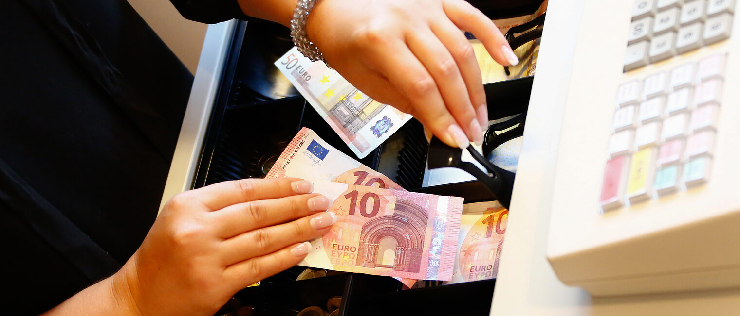 | Kasse | Bonpflicht | TSE | Kassen-Meldepflicht | Datensicherheit Nahaufnahme zweier Hände die Geld in eine Kasse einsortieren