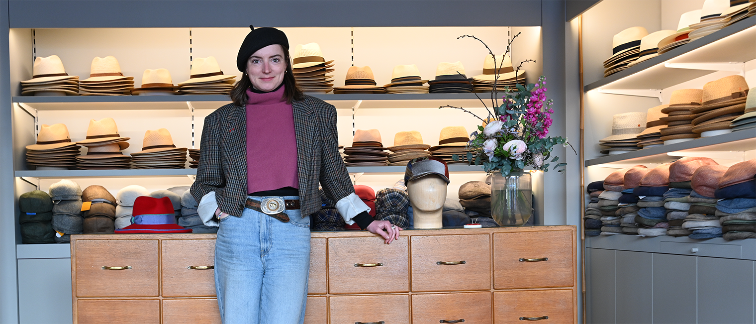 Modistin Laura Zieger Modistin Laura Zieger in ihrem Geschäft in Würzburg vor einer Kommode, auf der Hüte liegen; im Hintergrund Regale mit Hüten.