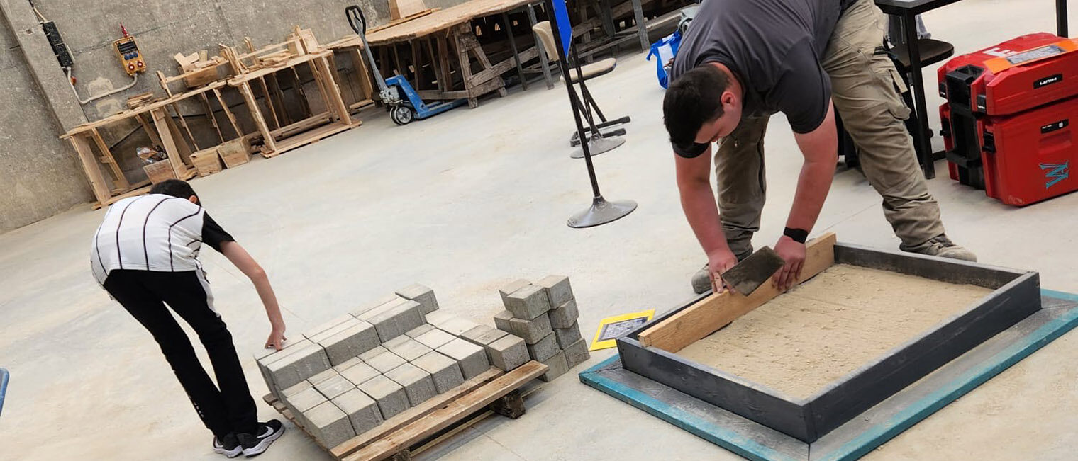 Jugendliche in der Halle der Bauinnung legen helle Pflastersteine auf Sand in einem Quadrat