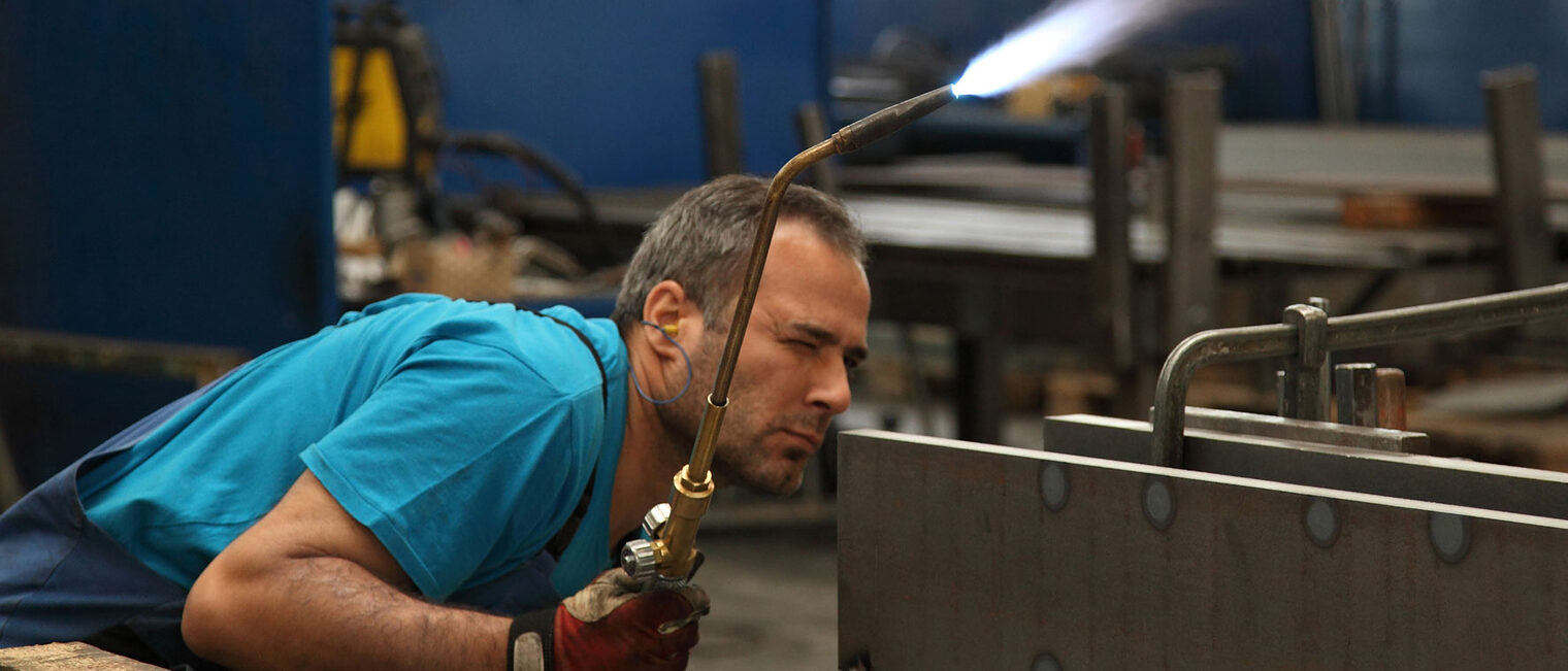 Metallbauer in Schutzkleidung mit Schweißgerät in der rechten Hand inspiziert gebückt die Schweißnaht 