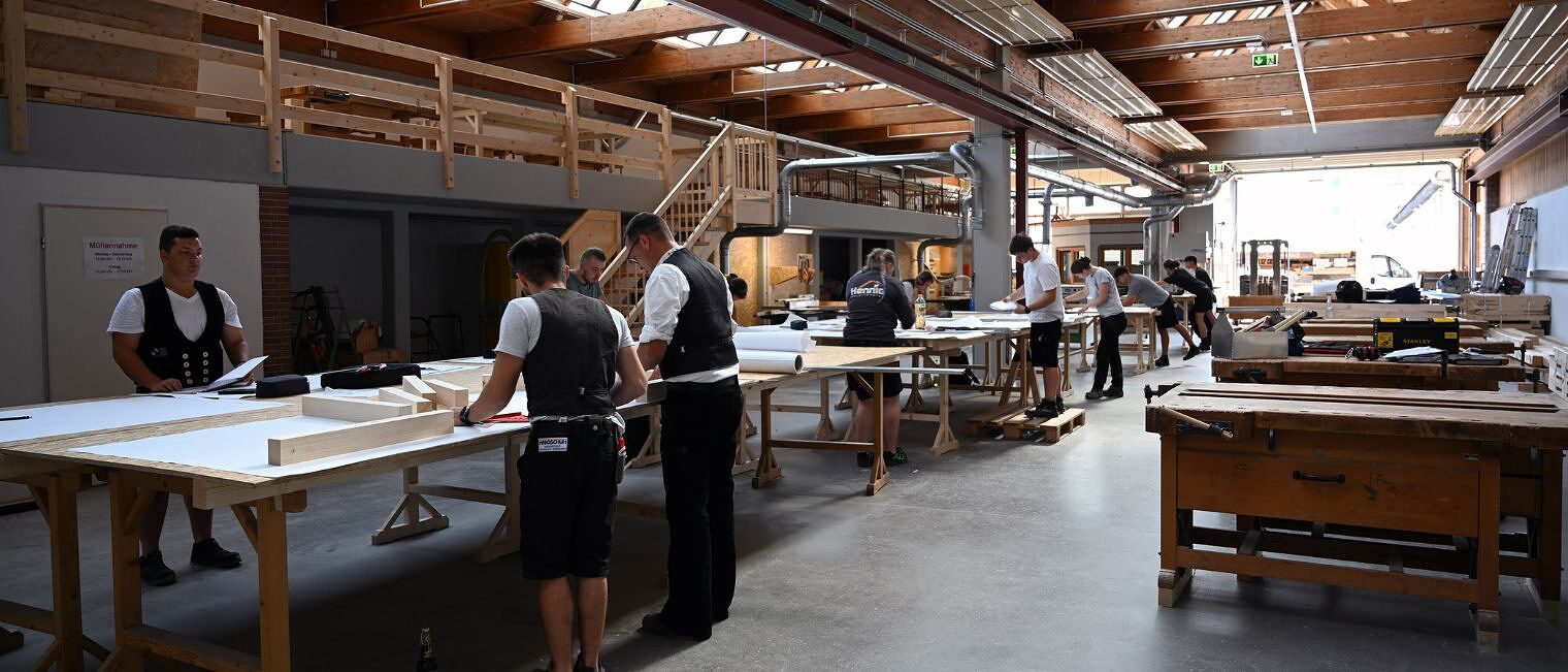 Große Halle mit mehreren Arbeitstischen, an denen Zimmerer-Meisterschüler in Arbeitskleidung und Kluft arbeiten