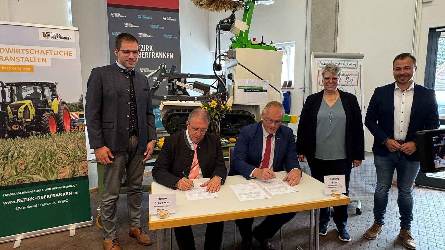 Fünf Personen sind auf dem Bild. Zwei sitzen am Tisch und unterschreiben einen Vertrag. Drei Personen stehendrumherum 