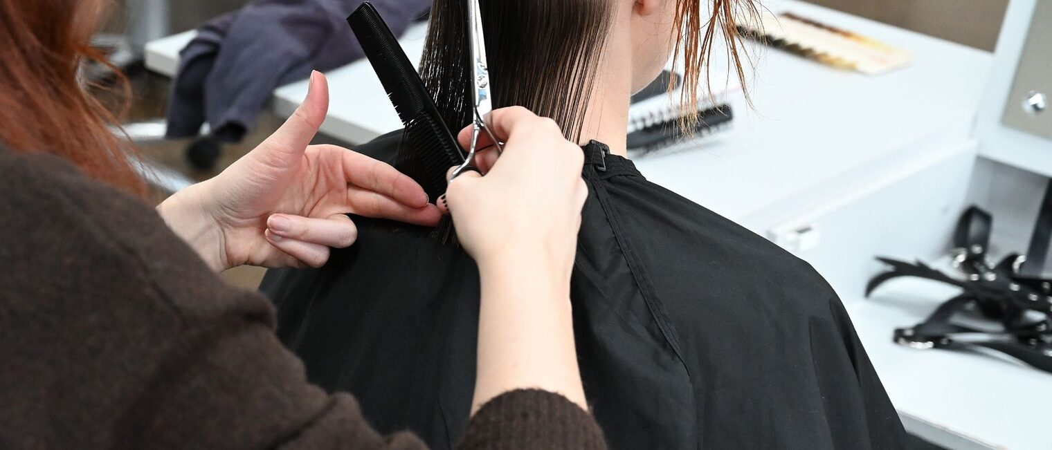 Situation von hinten fotografiert - eine Friseurin mit Kamm und Schere in der Hand schneidet einer sitzenden Dame die Haare